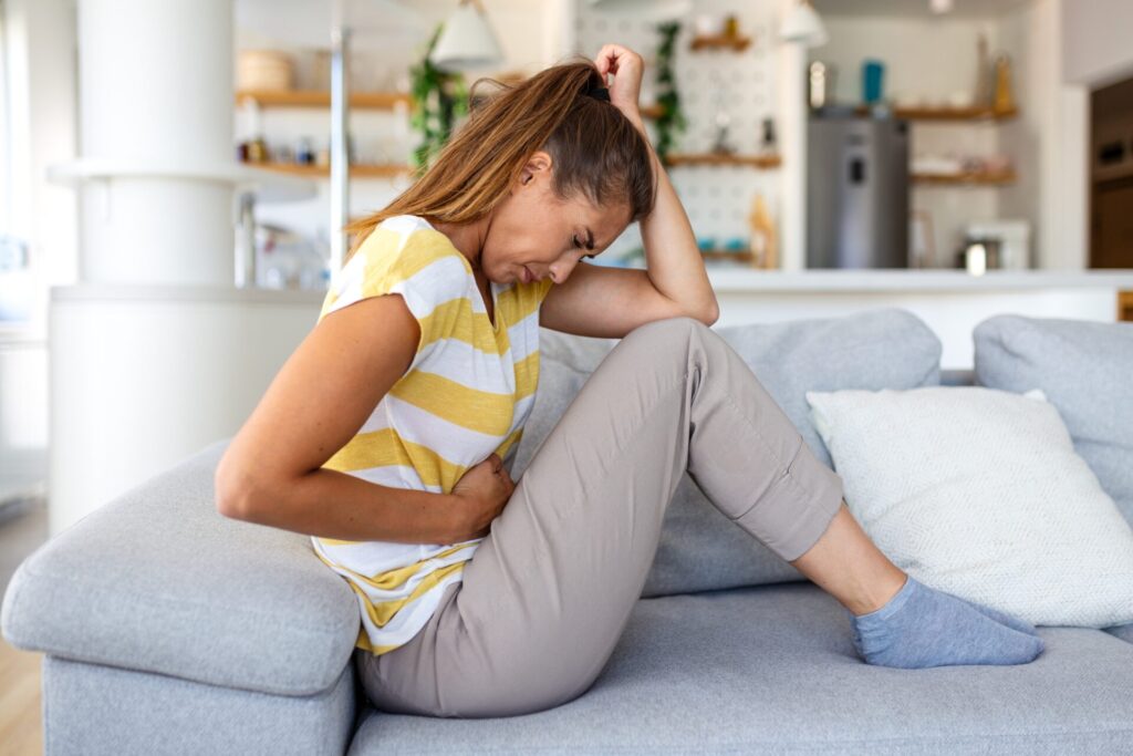 Woman,With,Stomach,Pain,Sitting,On,Sofa,Holding,Her,Belly.